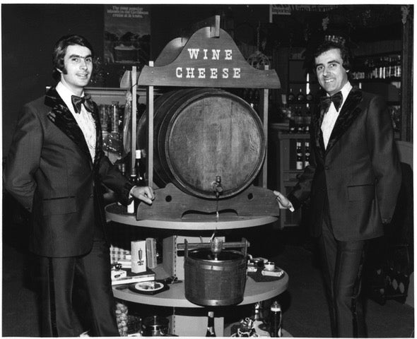 Two men in tuxedos standing beside a wine and cheese barrel display in a store, black-and-white photo.