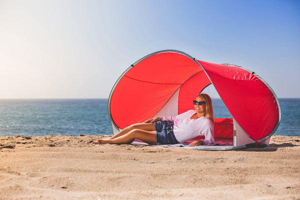 Woman relaxing under a red beach shade tent on sandy shore beside the ocean.