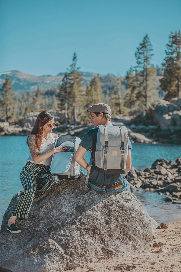 Two people sit on a rock by a lake; a woman unpacks a grey picnic backpack while a man with a backpack looks on.