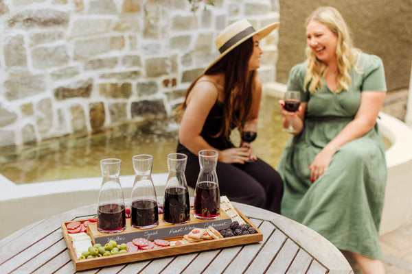Two women laugh with wine by a fountain; a table shows four labeled wine carafes and snacks.