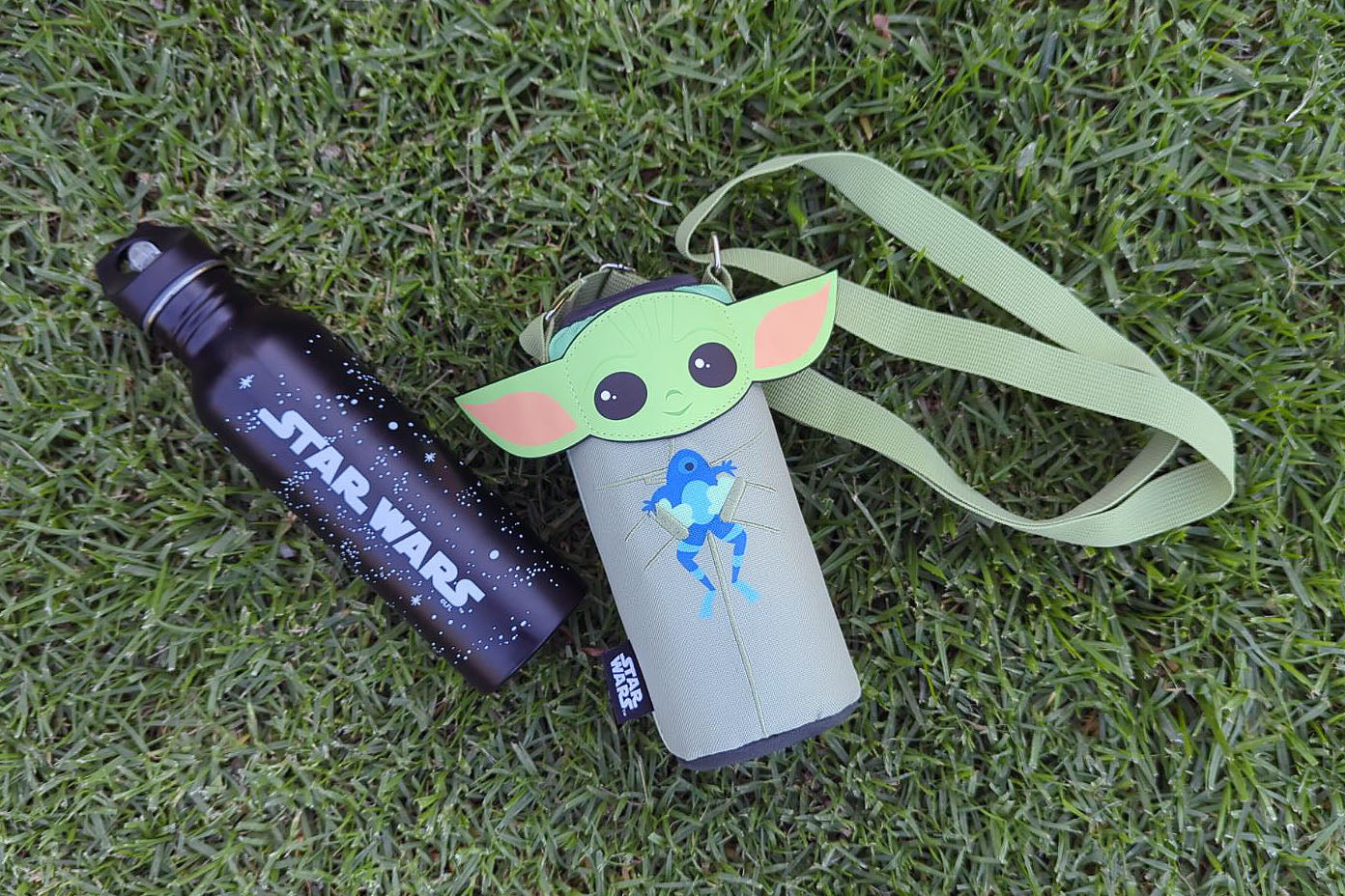 Star Wars water bottle and Grogu-themed shoulder pouch lying on green grass.