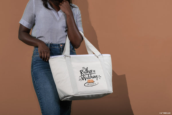 Person holding white tote bag with “I’d rather be watching Friends” text, wearing striped shirt and jeans