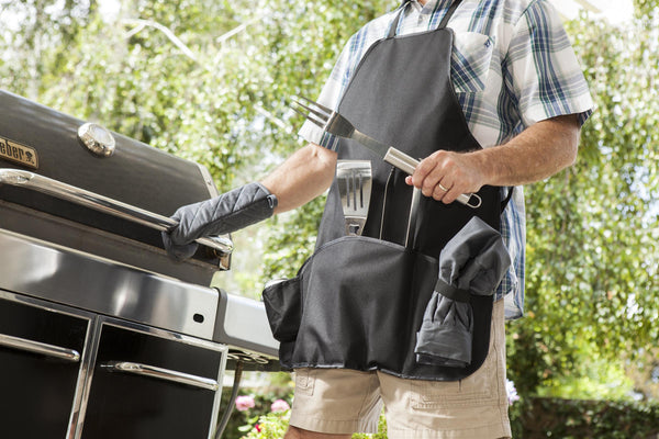 Person wearing a grilling apron and oven mitts beside a barbecue grill, holding BBQ tools outdoors.