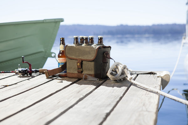 Fishing gear, beer six-pack in canvas bag, and rope on sunny wooden dock beside green boat on calm lake.