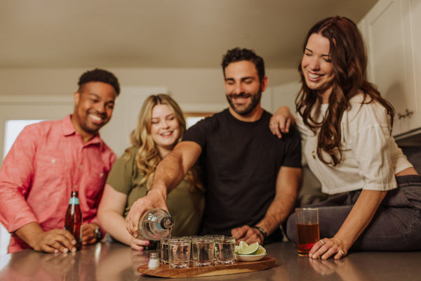 Four friends gathered around a kitchen counter, one pouring clear liquor into shot glasses on a tray with lime wedges.
