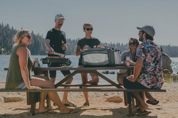 Group of friends enjoying drinks and grilling food at a lakeside picnic table with a cooler bag.