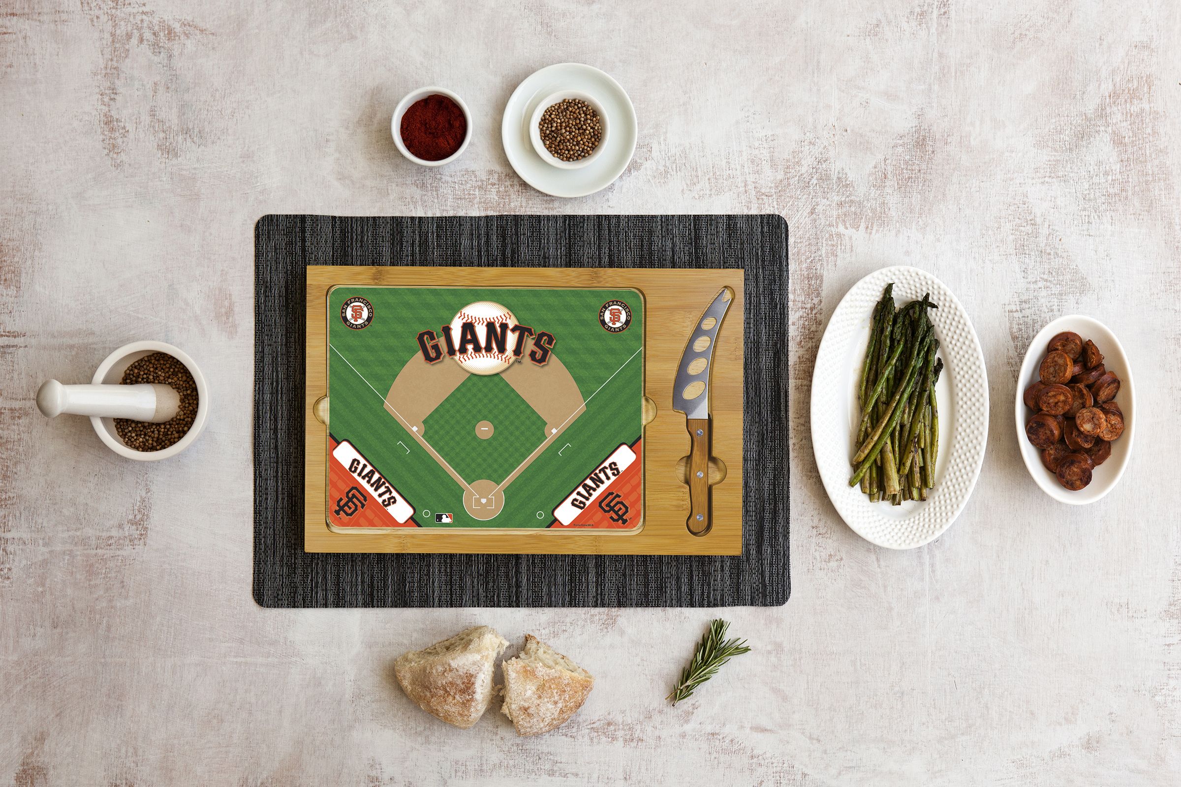 Baseball-themed cutting board with Giants field design, cheese knife, spices, bread, asparagus, and sausage sides.