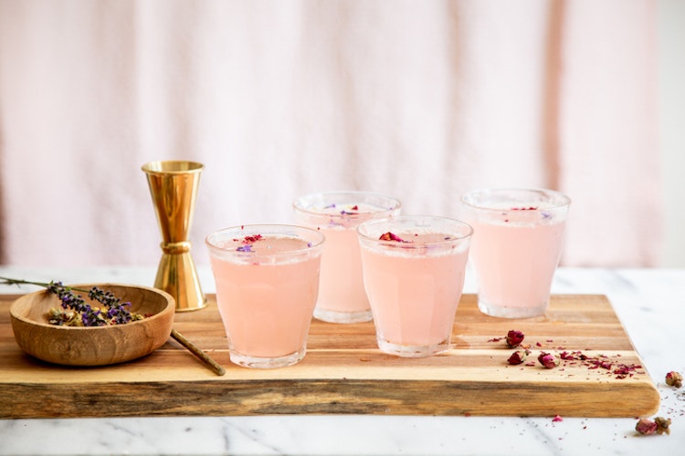 Four glasses of pink cocktails with rose petals on wooden board with jigger and dried lavender.
