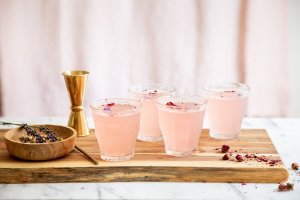 Four glasses of pink cocktails with rose petals on wooden board with jigger and dried lavender.