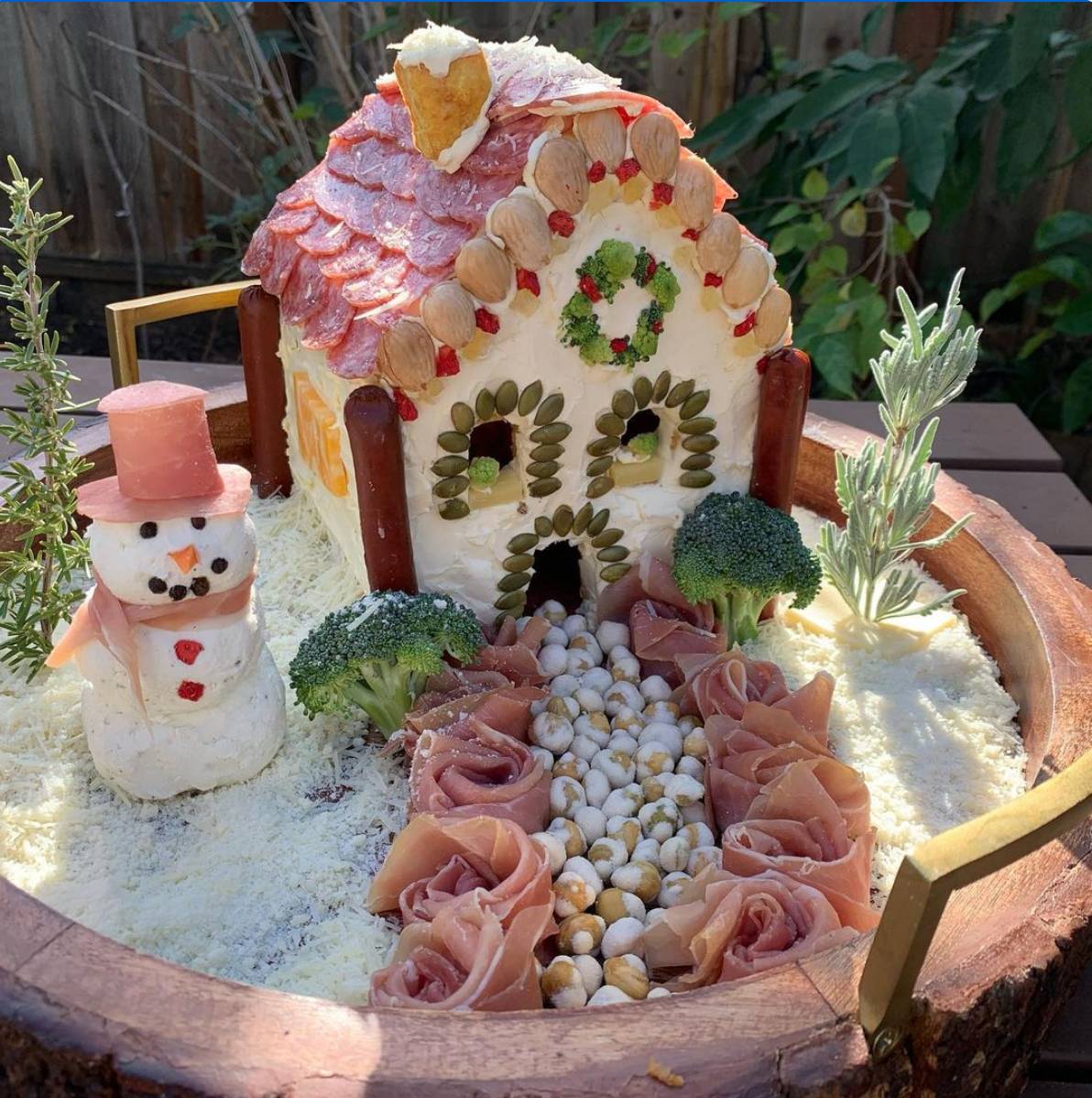 Whimsical cheese-and-meat house on wooden platter, salami roof, almond details, broccoli trees, cheese snowman.