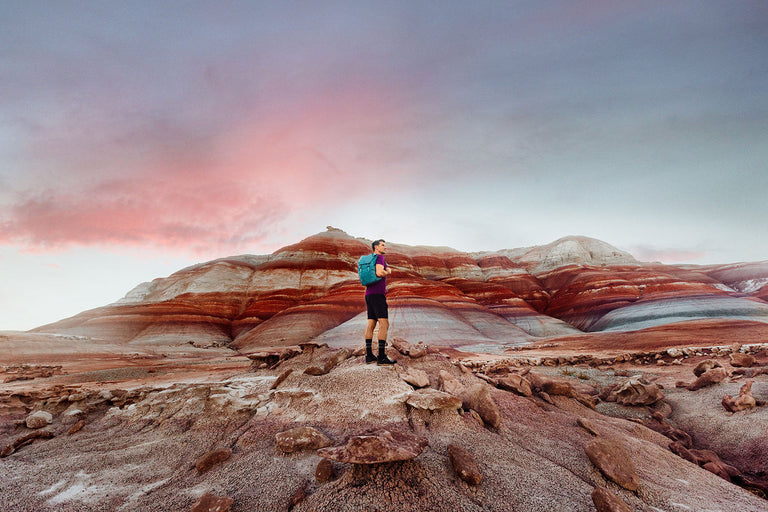 Hiker with backpack standing on rocky desert landscape beneath colorful striped hills and sky.