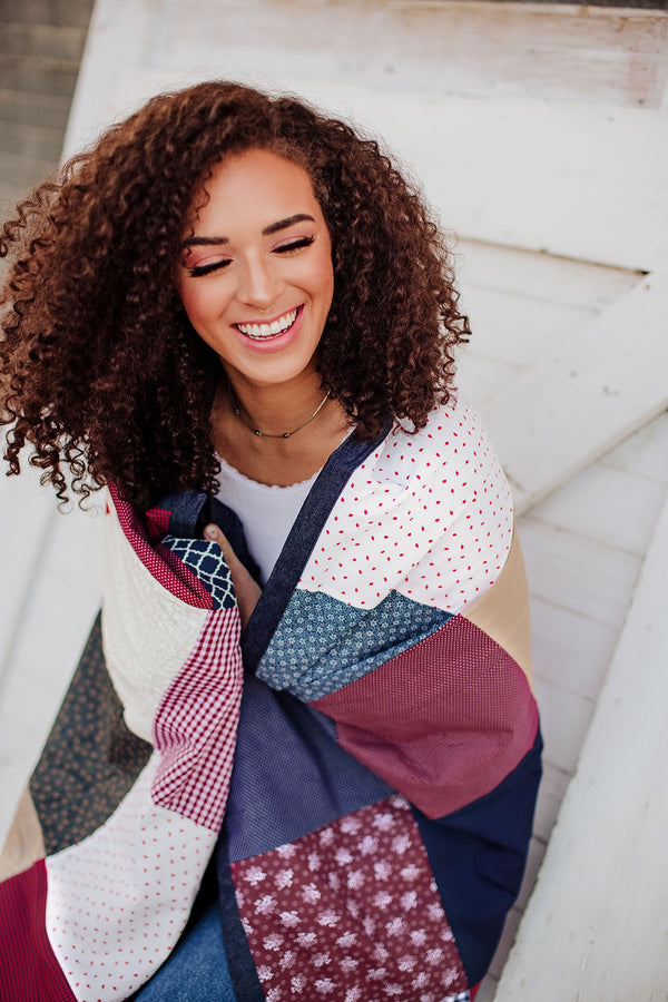 Smiling woman with curly hair wrapped in a colorful patchwork quilt, sitting on outdoor steps.