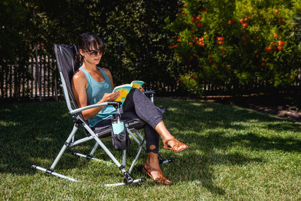 Woman relaxing on lawn chair in sunny garden, reading book, wearing sunglasses, tank top and leggings, peaceful scene.