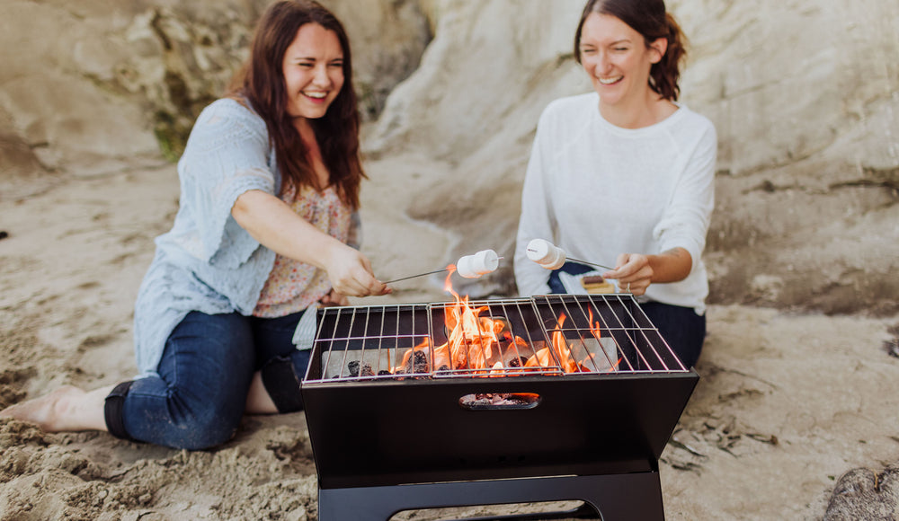 two woman making s'mores at the beach with portable folding charcoal grill