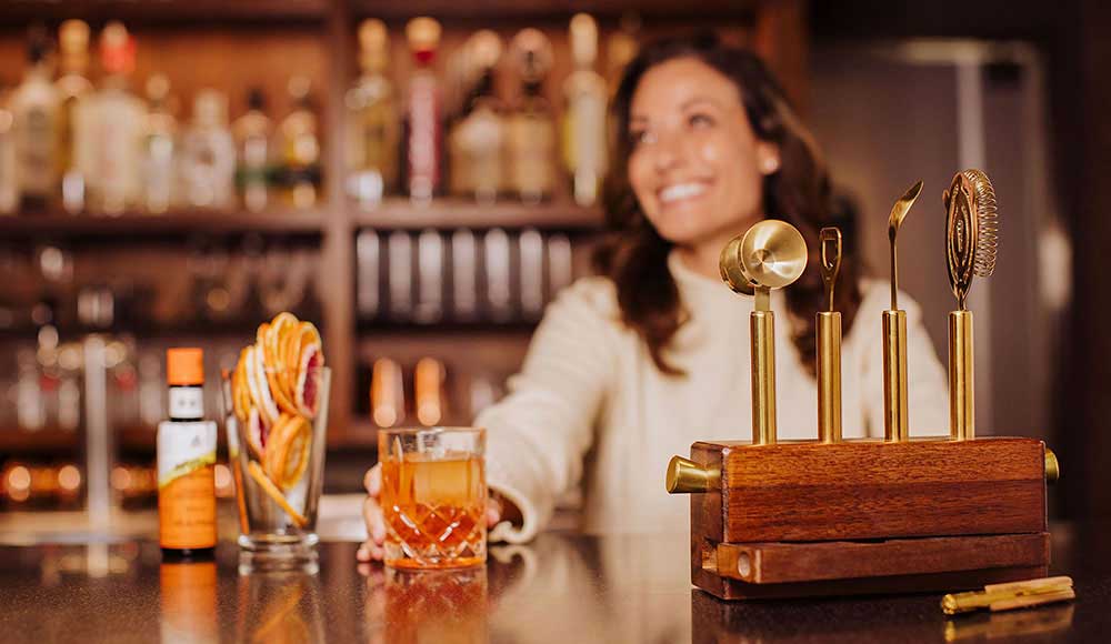 Holding a craft cocktail on a bar top with a wood and gold tone cocktail making set in the foreground