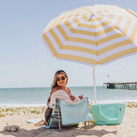 Person smiling on the beach in an outdoor chair under a yellow and white striped umbrella with a tote bag nearby