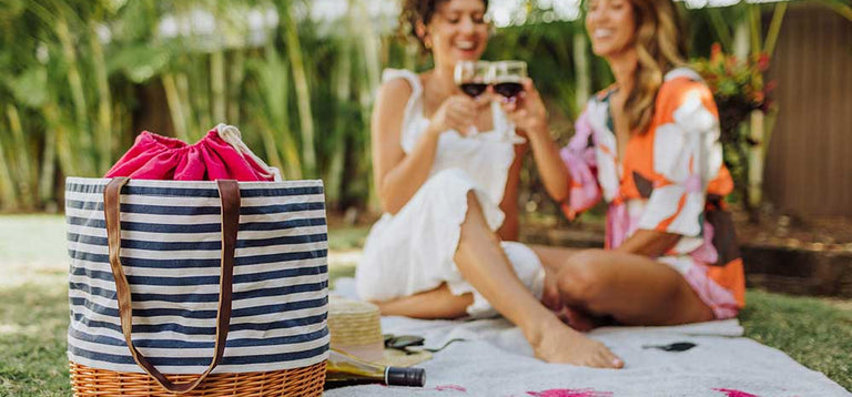 Two people sitting barefoot on a picnic blanket and toasting with red wine glasses with a striped Picnic Time tote in the foreground